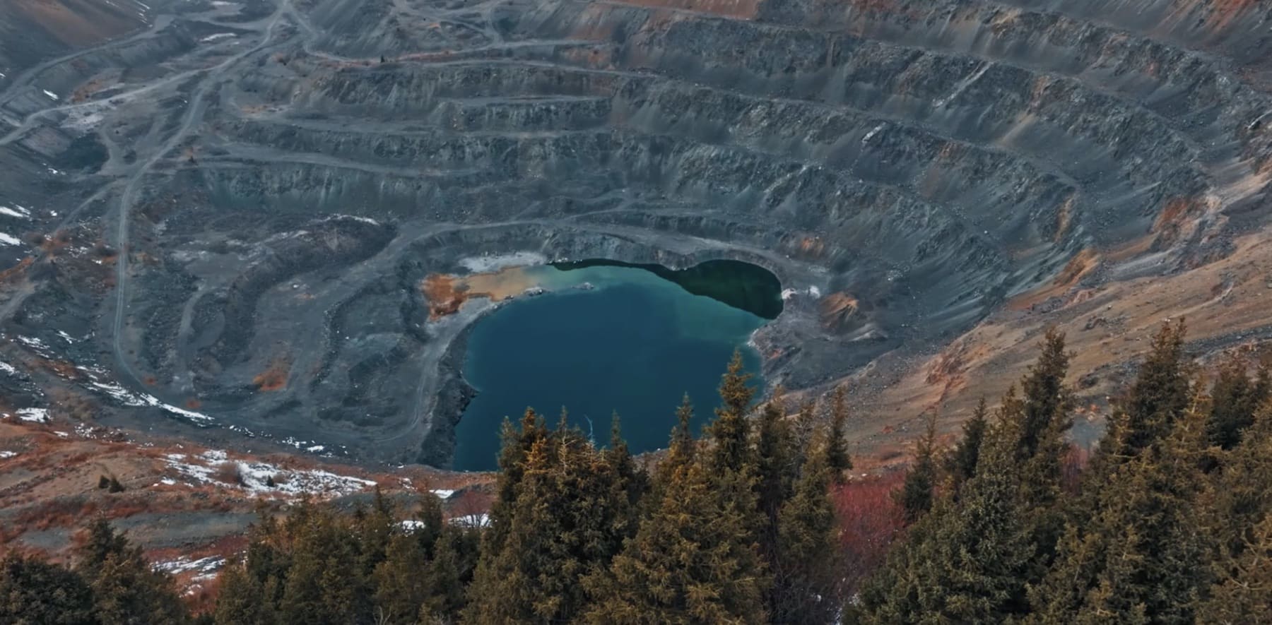 Aerial view of an open pit mine filled with blue water, surrounded by terraced rocky slopes and evergreen trees in the foreground.